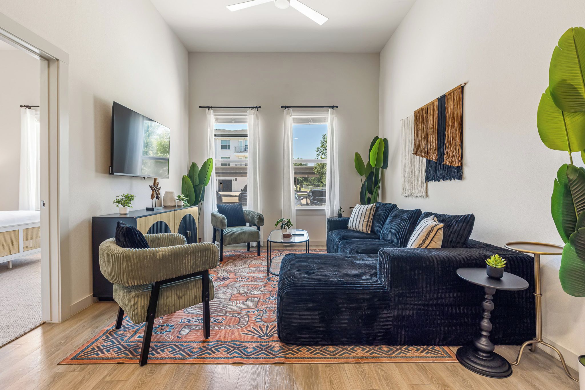 A living room with a sofa, armchairs, a coffee table, and a TV. There are large windows with curtains, a patterned rug, and a plant.