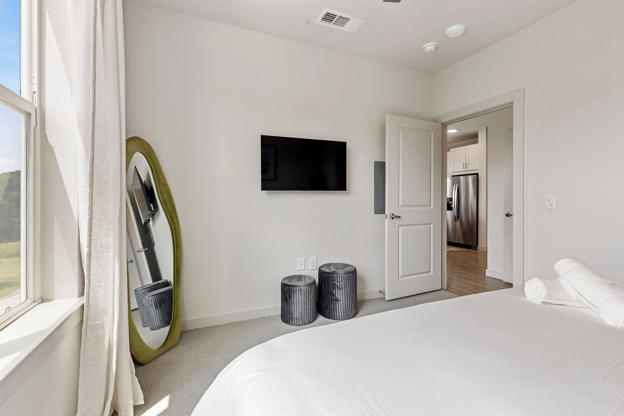 A modern bedroom with a large bed, a television mounted on the wall, and a door leading to a kitchen.