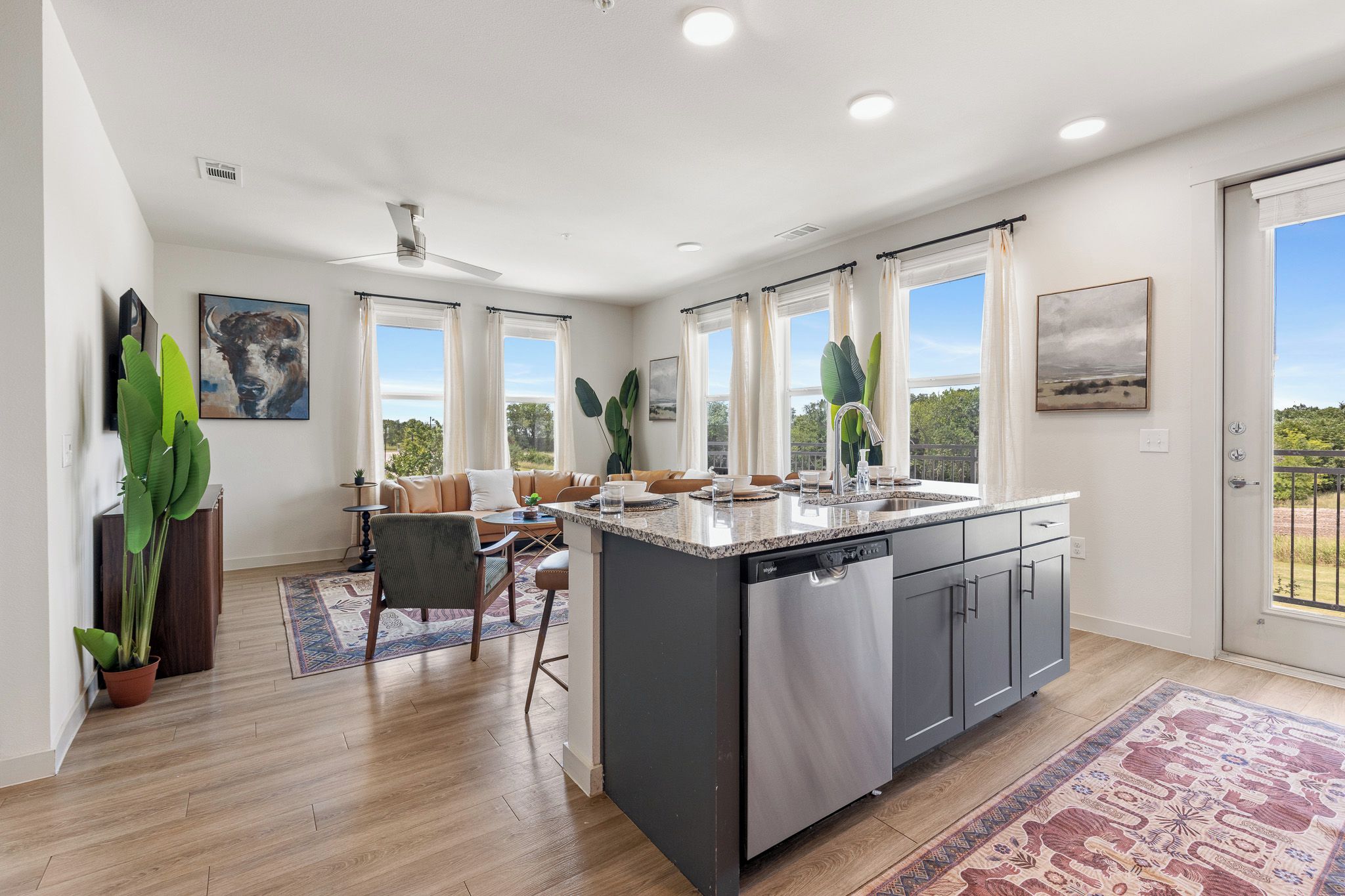 A modern kitchen with a central island, a dining area with a sofa and chairs, and large windows with curtains.