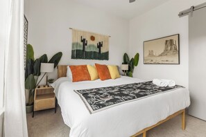 Un dormitorio con cama, almohadas, una mesita de noche, lámpara, un adorno en la pared y un cuadro enmarcado.