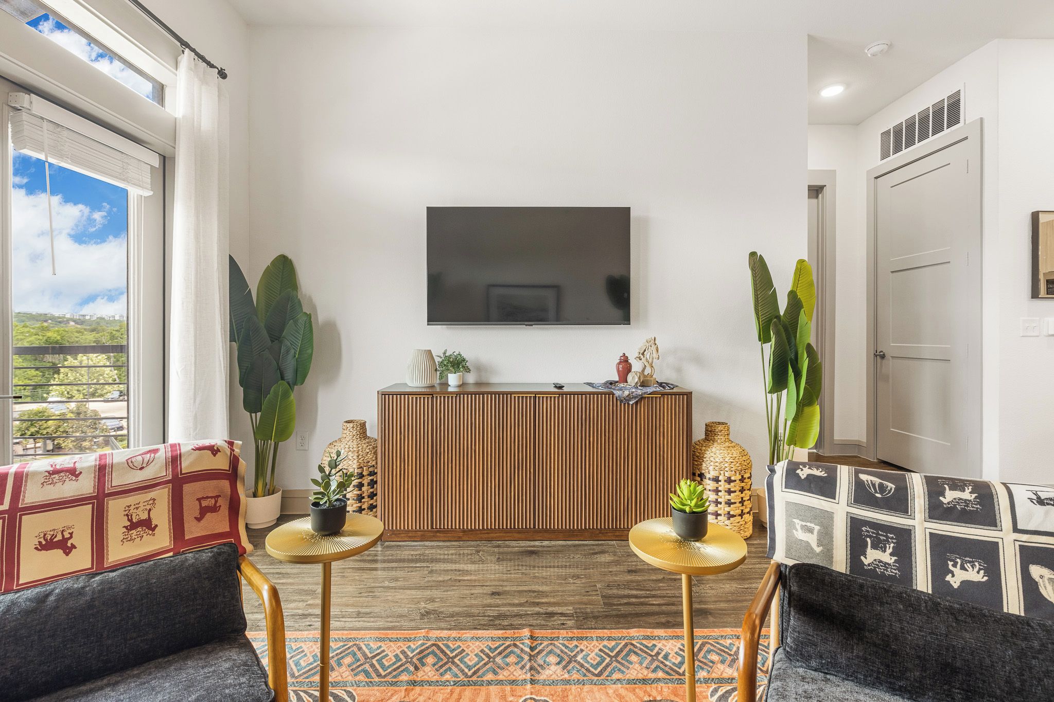 Una sala moderna con un televisor de pantalla plana montado en la pared, un mueble de madera, dos mesas redondas con plantas en macetas y una alfombra estampada.