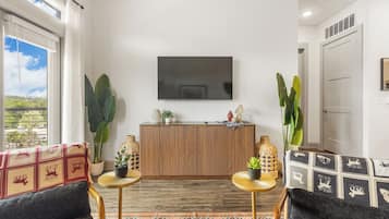 Una sala moderna con un televisor de pantalla plana montado en la pared, un mueble de madera, dos mesas redondas con plantas en macetas y una alfombra estampada.