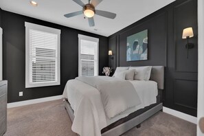 Ein Schlafzimmer mit einem Bett, zwei Fenstern mit Jalousien, einem Deckenventilator, einer Wandleuchte und einem Gemälde.