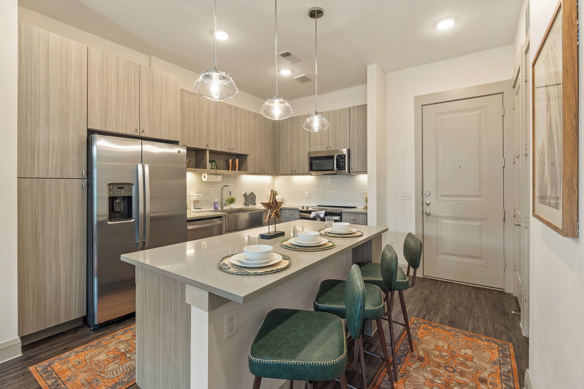 A modern kitchen with a central island, stainless steel appliances, pendant lighting, and a patterned rug.