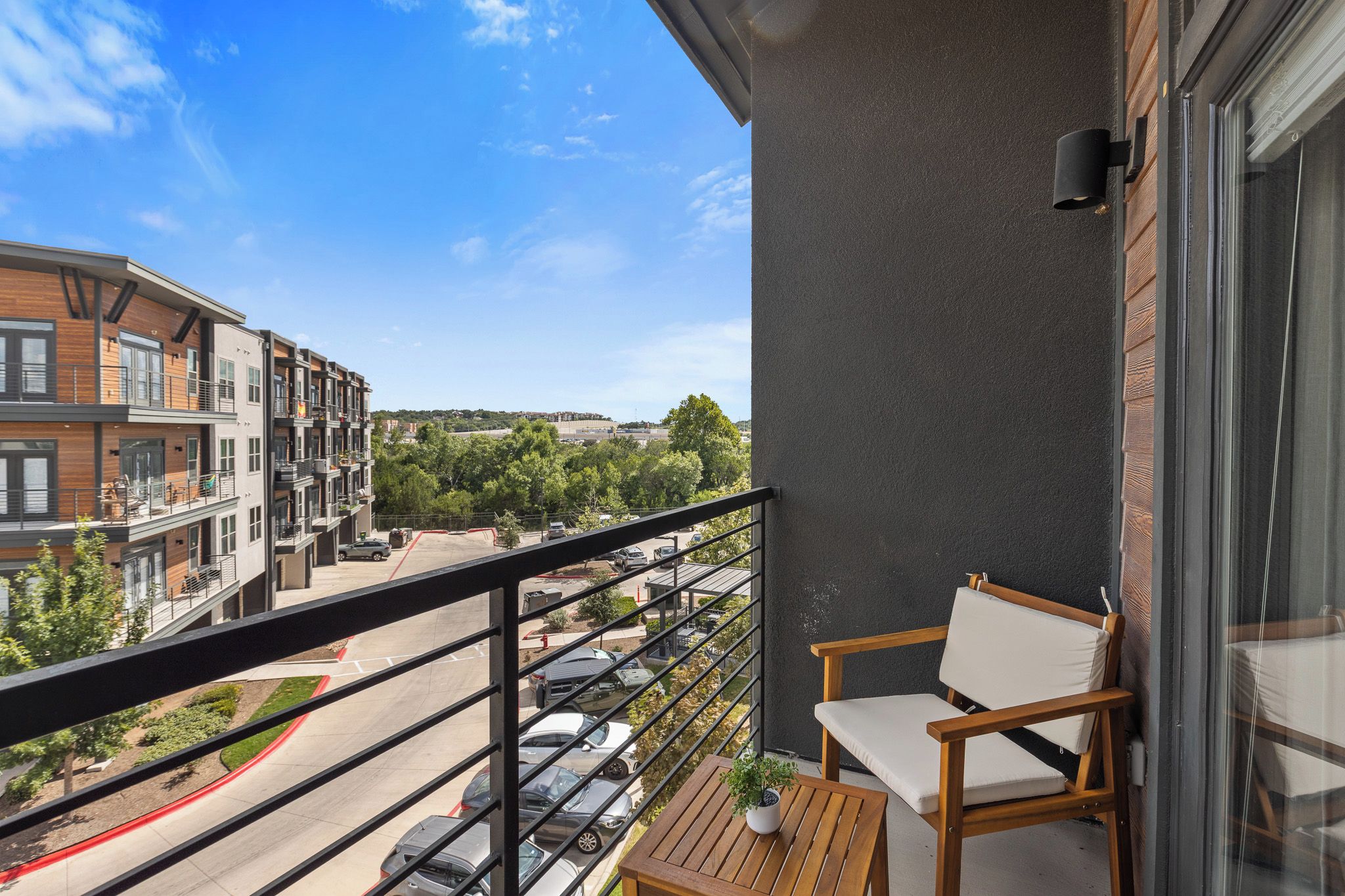 Een balkon met uitzicht op een parkeerplaats en gebouwen, een houten tafel en twee stoelen.