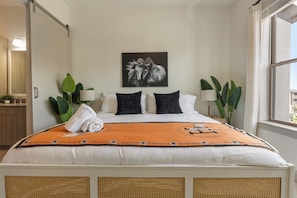 Een net opgemaakt bed met een geborduurd plaid, een handdoek en twee kussens. Rechts een groot raam en links een deur.
