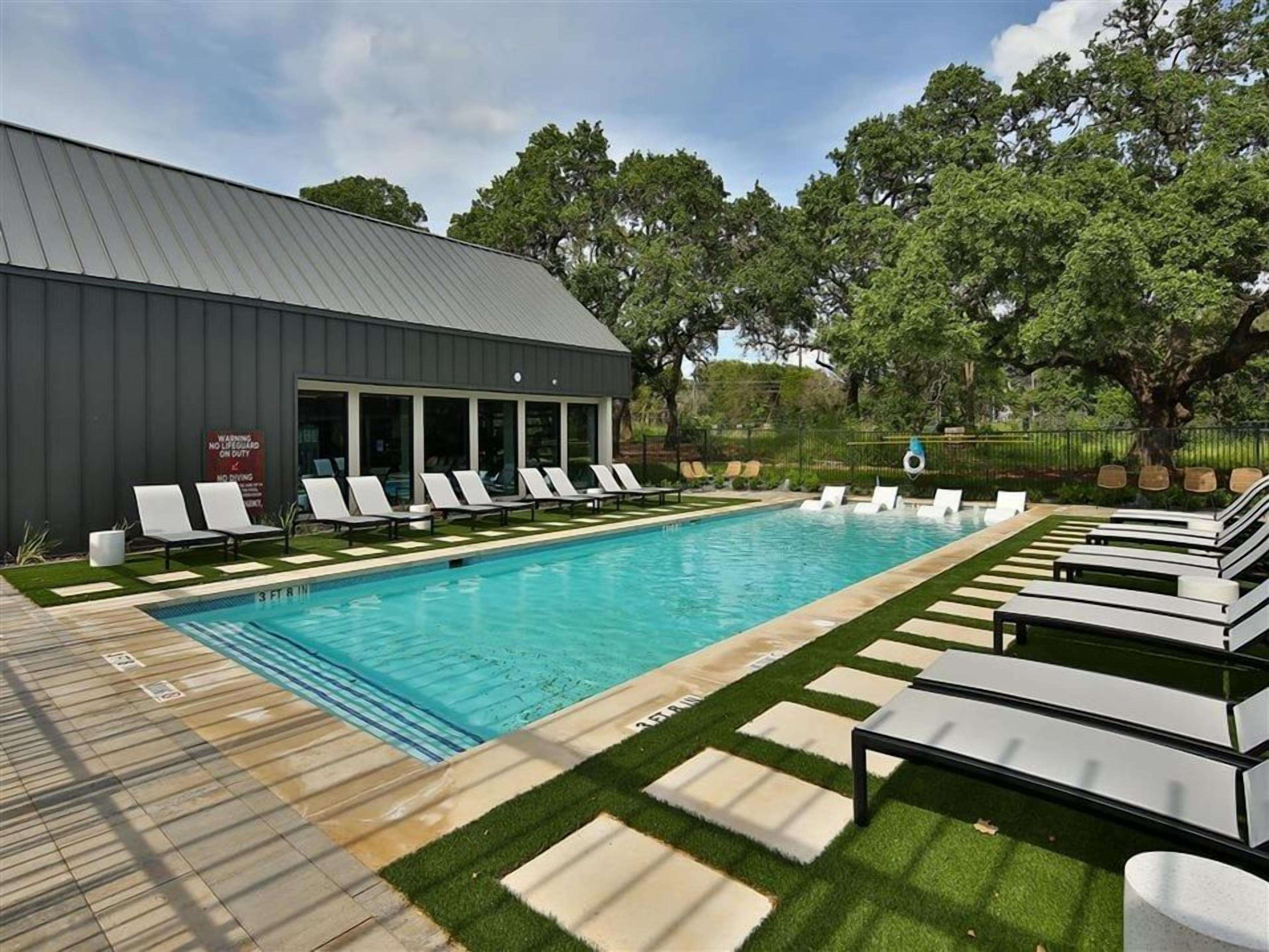 Une piscine avec des chaises longues, un bâtiment doté de grandes fenêtres et une zone herbeuse.