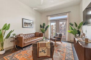 Een woonkamer met een bruine leren bank, een kamerbreed tapijt, een salontafel en een fauteuil. Er is een groot raam met gordijnen, een ingelijst schilderij aan de muur en een potplant.