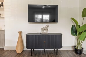 Ett modernt vardagsrum med en platt-TV som är monterad på väggen, ett mörkt konsolbord med en dekorativ hästskulptur, en korgvas och en stor grön växt i en svart kruka.