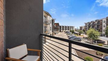 Un balcon avec une chaise en bois, une vue sur un parking et, au loin, des immeubles à plusieurs étages.