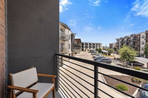 Un balcon avec une chaise en bois, une vue sur un parking et, au loin, des immeubles à plusieurs étages.