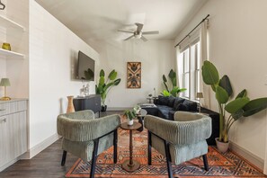 Un salon comprenant un canapé, deux fauteuils, une table basse et un téléviseur fixé au mur. On y trouve également un ventilateur de plafond, une fenêtre avec des rideaux et un tapis à motifs.