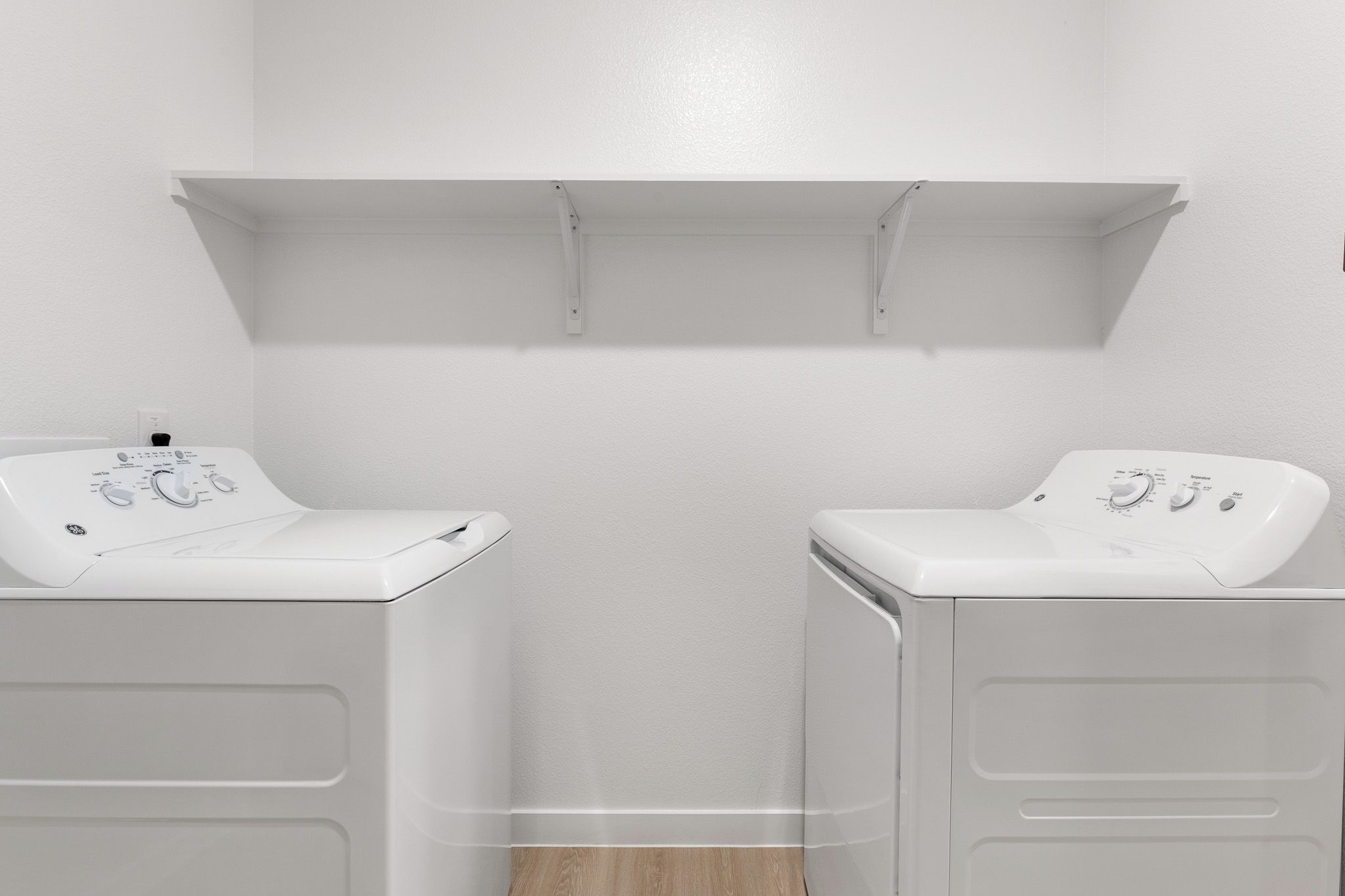 A laundry room with two white washing machines and a white shelf above them.