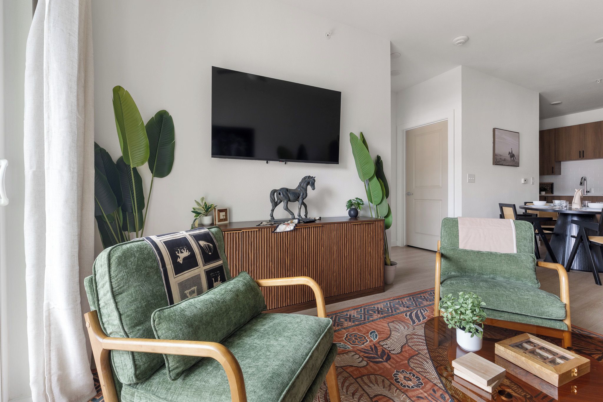 A modern living room with a flat-screen TV, a wooden entertainment unit, green upholstered chairs, a patterned rug, and a dining area with a table and chairs.