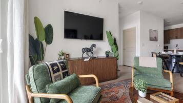 A modern living room with a flat-screen TV, a wooden entertainment unit, green upholstered chairs, a patterned rug, and a dining area with a table and chairs.