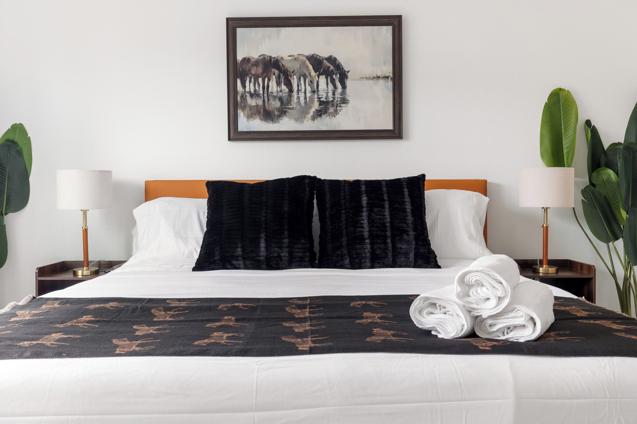 A hotel room with a bed, two nightstands, a painting, and potted plants.