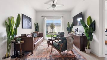 Ein Wohnzimmer mit einer Couch, Sesseln, einem Couchtisch, großen Kübelpflanzen, einem Deckenventilator und einem an der Wand befestigten Fernseher.