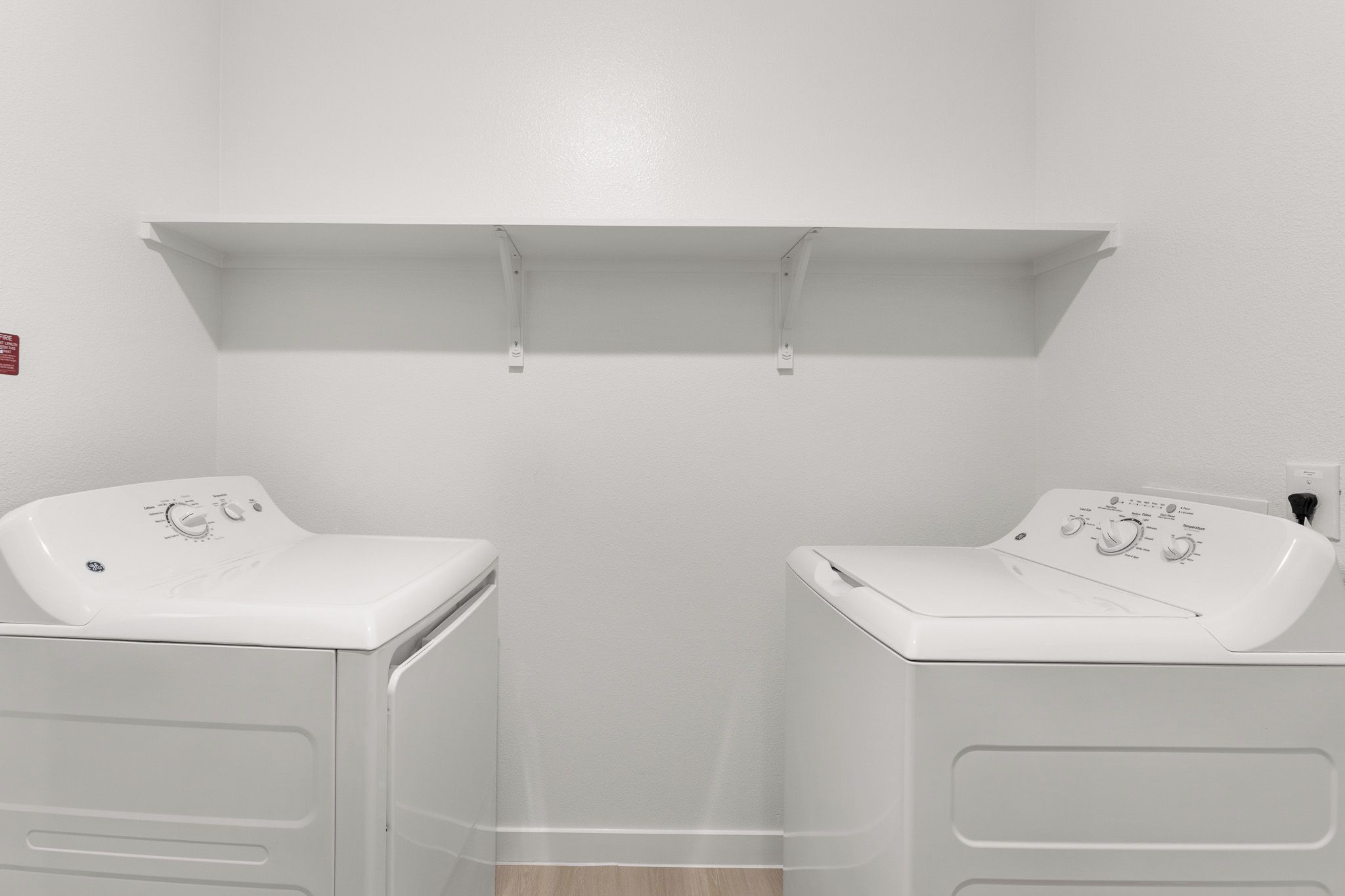 A laundry room with two white washing machines, a shelf above, and a fire alarm on the wall.