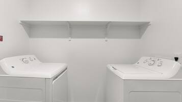 A laundry room with two white washing machines, a shelf above, and a fire alarm on the wall.