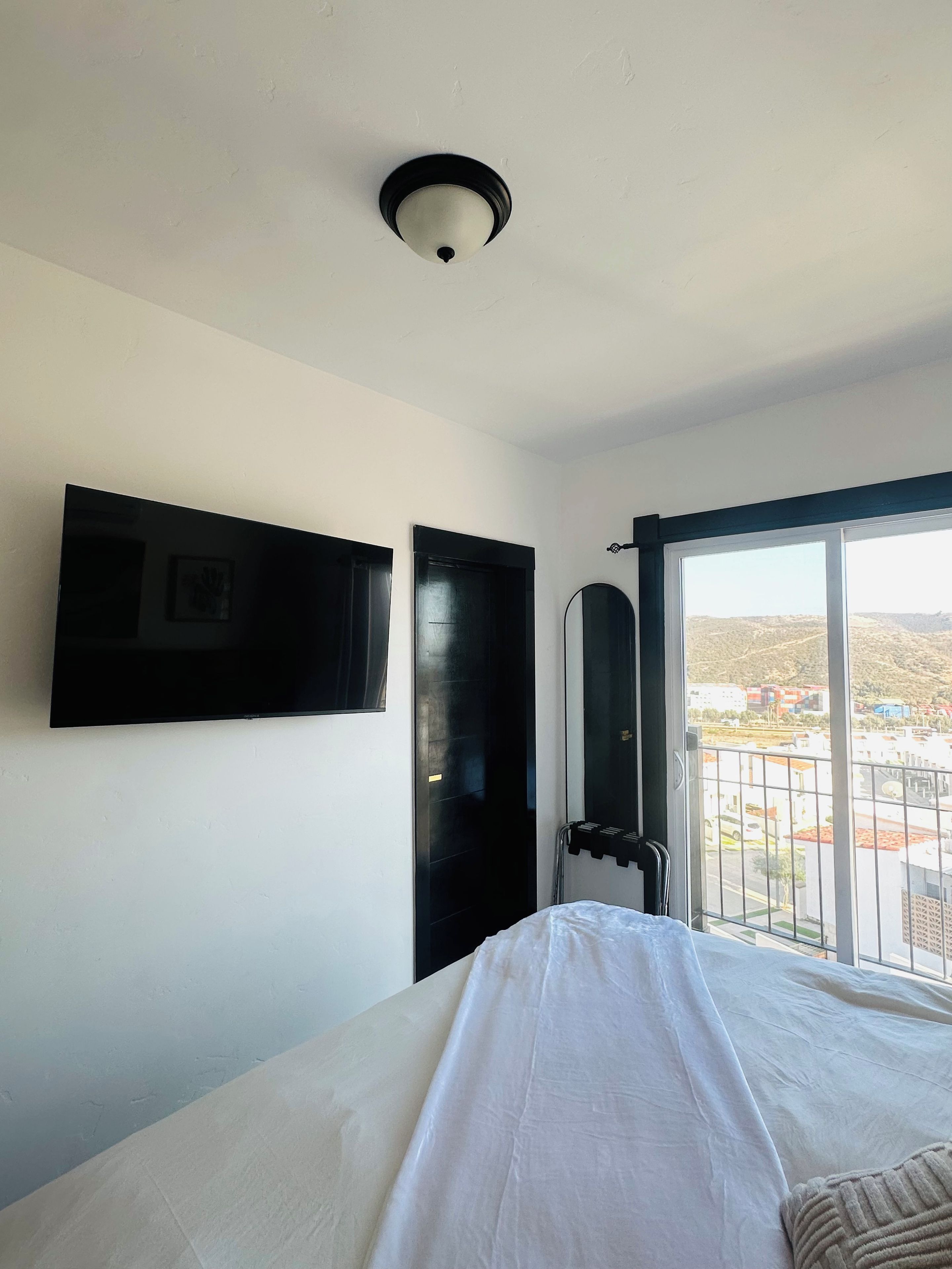 A hotel room with a bed, a flat-screen TV mounted on the wall, a door, and a window with a view of buildings and hills.