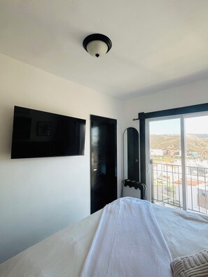 Une chambre d’hôtel équipée d’un lit, d’un téléviseur à écran plat fixé au mur, d’une porte et d’une fenêtre donnant sur des bâtiments et des collines.