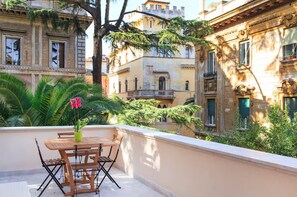 Ein Balkon mit Holztisch und Stühlen, umgeben von üppiger Vegetation und historischen Gebäuden.