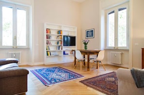 Ein Wohnzimmer mit einer Couch, einem Esstisch mit Stühlen, einem Bücherregal, einem Fernseher und einem Fenster.
