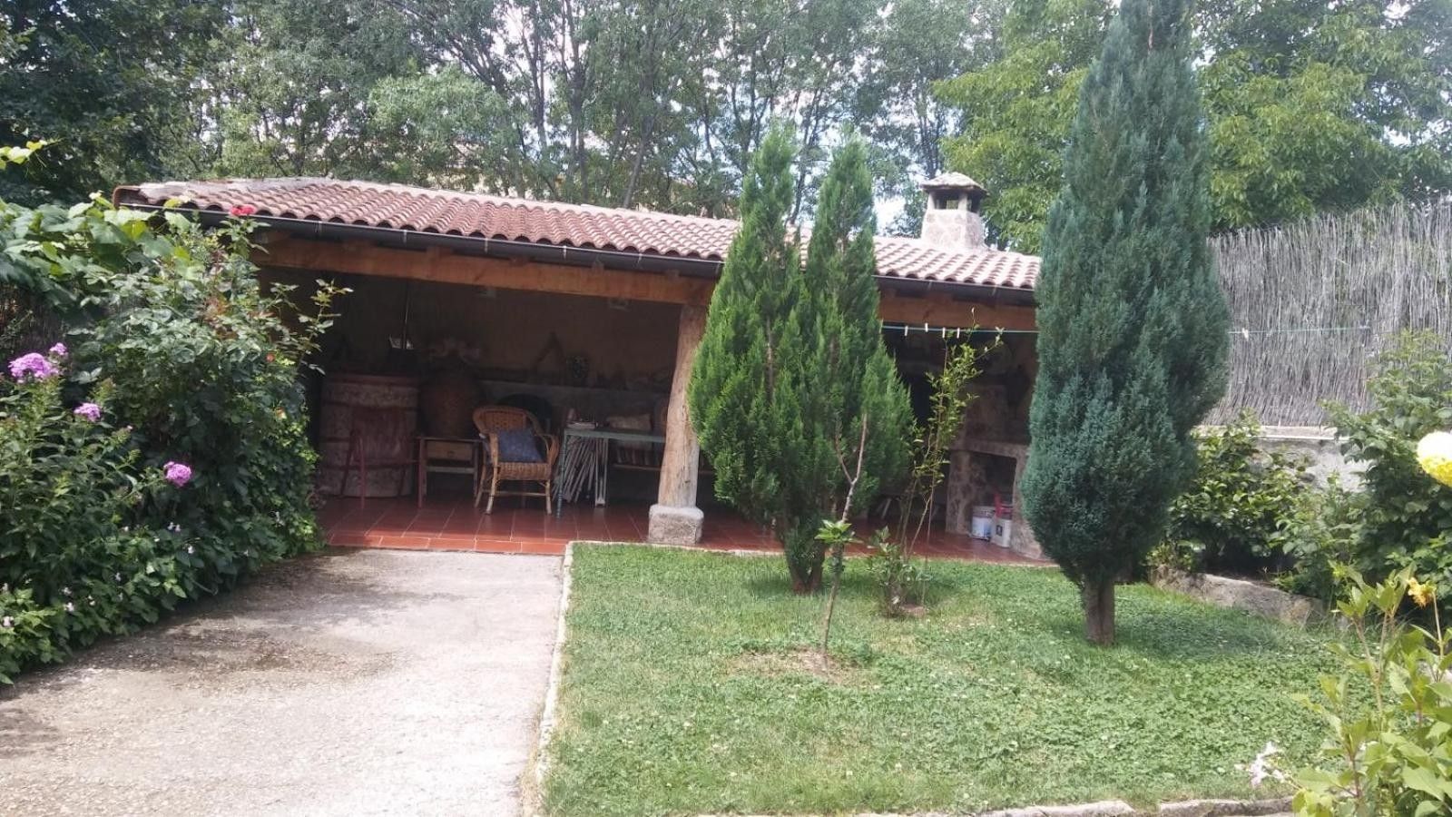 Ein rustikales Gebäude mit Ziegeldach, einer überdachten Terrasse mit Gartenmöbeln und einem gepflegten Garten mit verschiedenen Pflanzen und Rasenfläche.