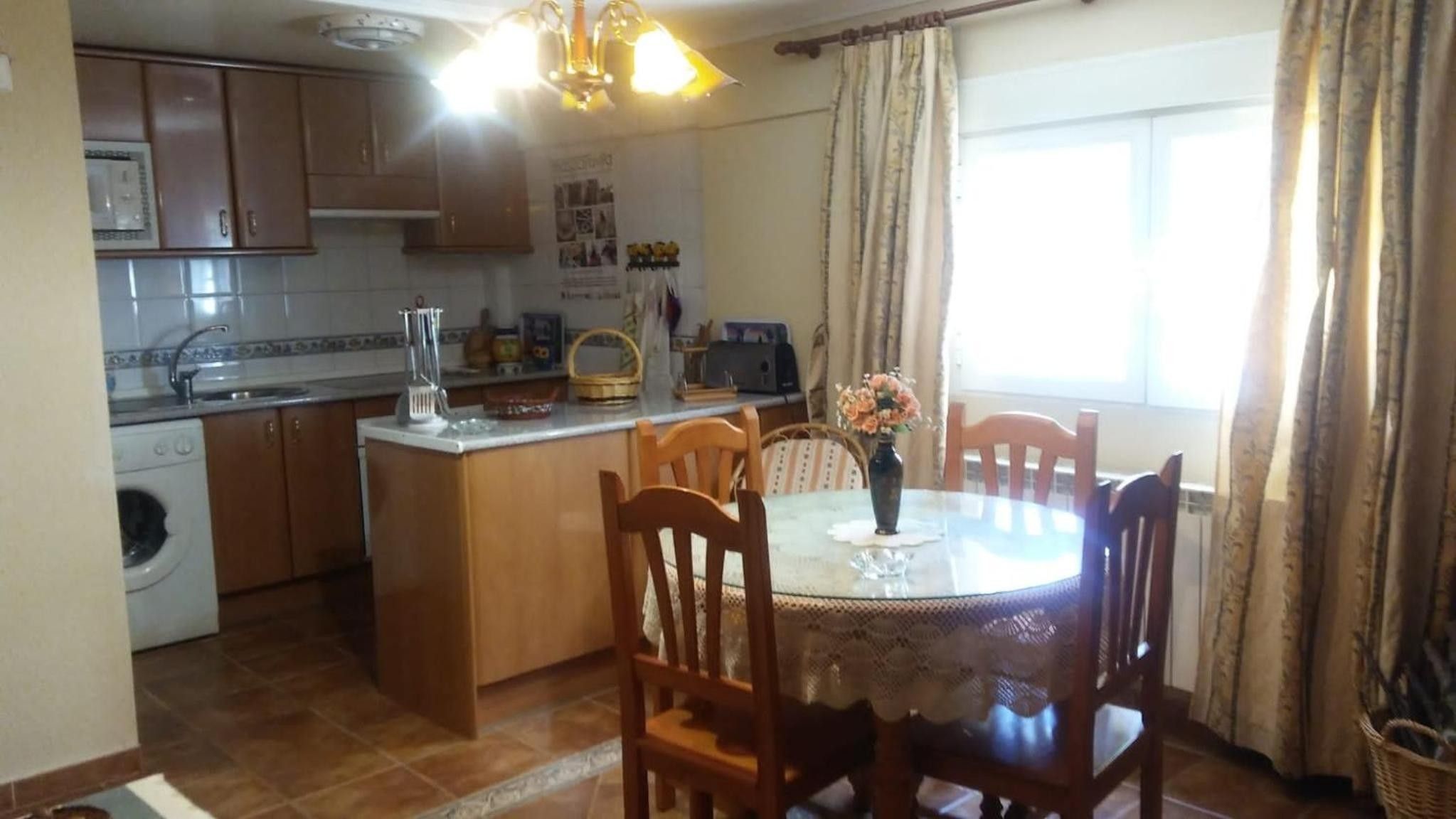 Una cocina con armarios de madera, lavadora, mesa de comedor con sillas y ventana con cortinas.