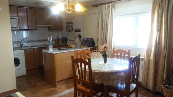 A kitchen with wooden cabinets, a washing machine, a dining table with chairs, and a window with curtains.