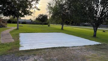 Un área pavimentada rodeada de césped y árboles, con una acera y una valla al fondo.