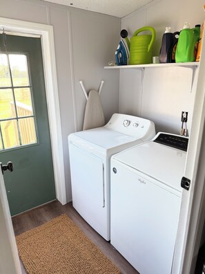 A laundry room with a front-loading washer and dryer, a drying rack, and a shelf with cleaning supplies. - The Cozy Olive Retreat – Peaceful Country Living – 3 Bedroom, 2 Bath (Santa Fe)