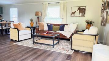 A living room with a brown leather sofa, two wicker armchairs, a glass coffee table, and a dining area with a wooden table and chairs.
