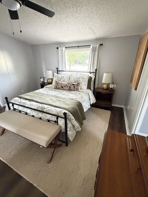 A bedroom with a bed, bedside tables, a bench, a ceiling fan, and a window with curtains.