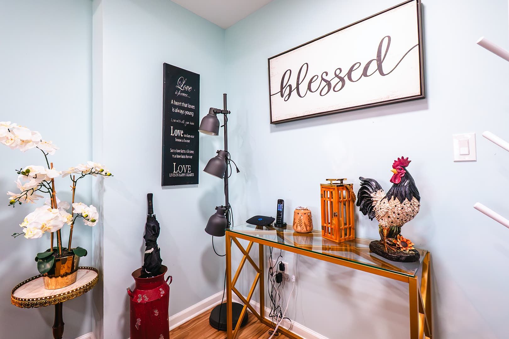 Une pièce avec un mur bleu clair, un panneau encadré portant l’inscription « béni », un coq décoratif, une petite table avec une lampe et un luminaire fixé au mur.