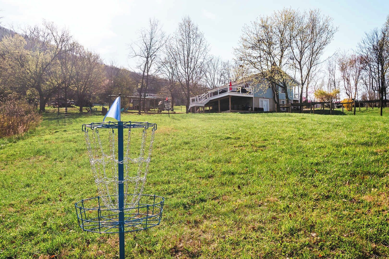 Una canasta de disc golf en un campo cubierto de césped, con una casa y árboles al fondo.