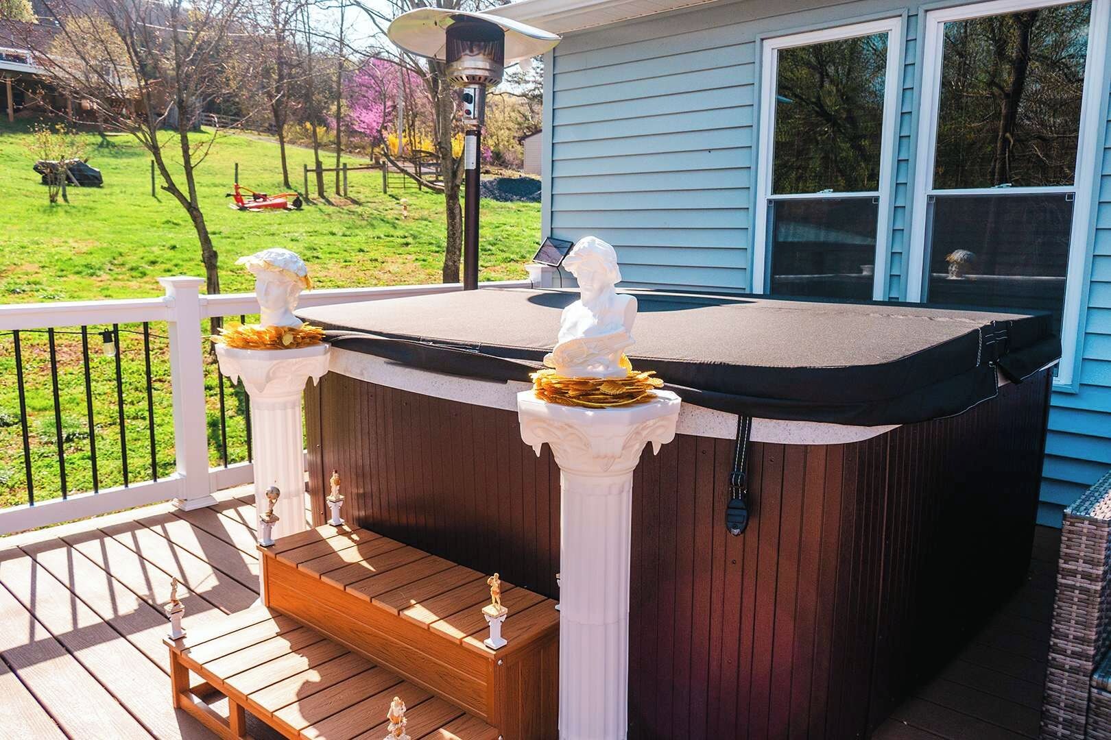 Un jacuzzi en una terraza de madera con barandillas blancas, una pequeña estatua y vistas a un jardín.
