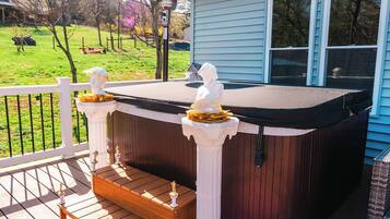 Un jacuzzi en una terraza de madera con barandillas blancas, una pequeña estatua y vistas a un jardín.
