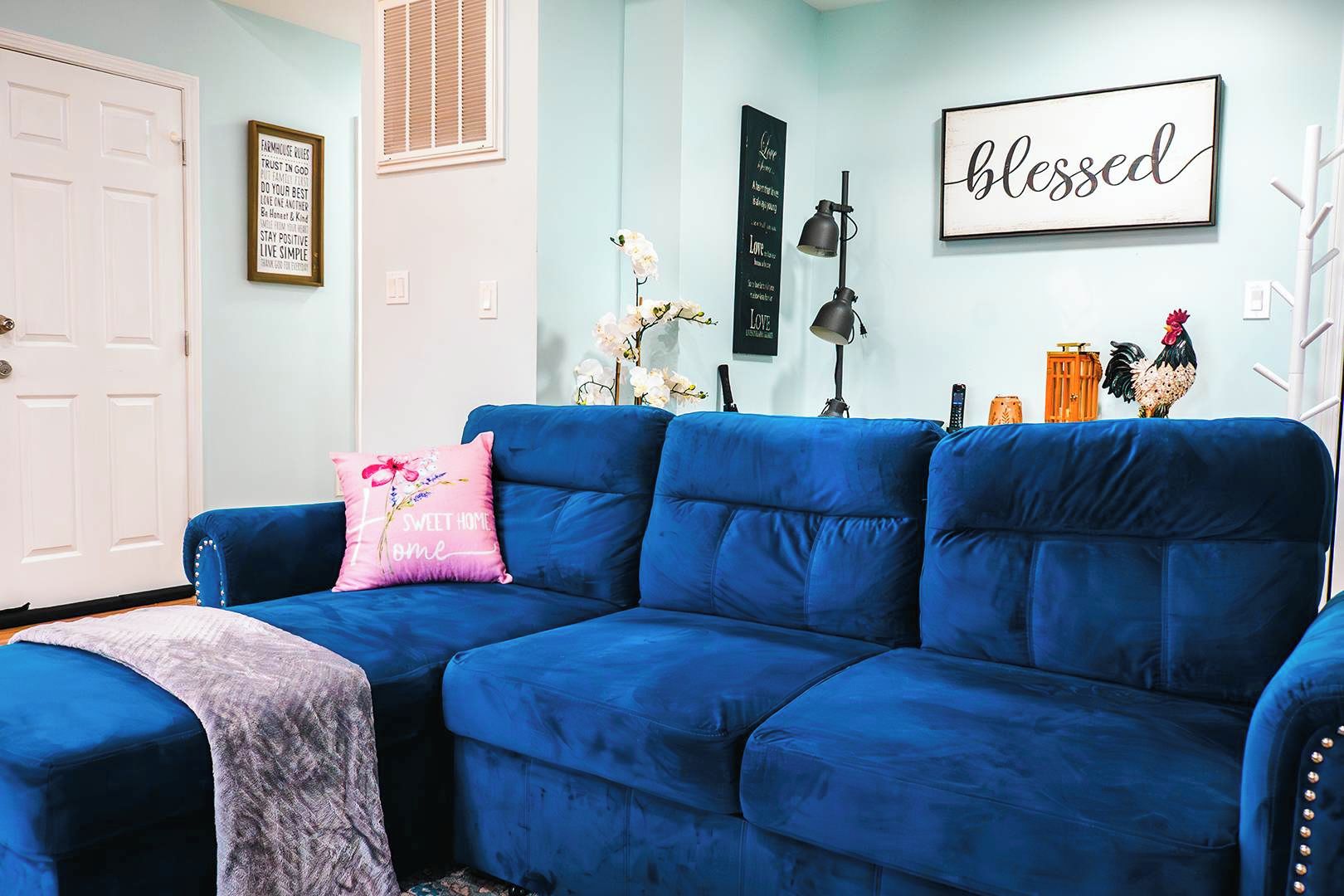 Um sofá azul com uma almofada rosa, uma porta branca e uma parede com frases motivacionais e uma placa escrita 'abençoado'.