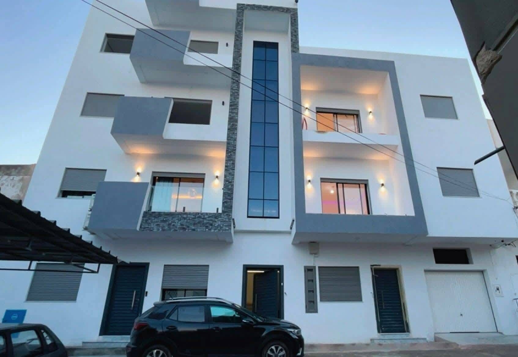 A multi-story building with a modern design, featuring a central staircase, multiple windows with external lighting, and a car parked in front.