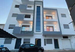 A multi-story building with a modern design, featuring a central staircase, multiple windows with external lighting, and a car parked in front.