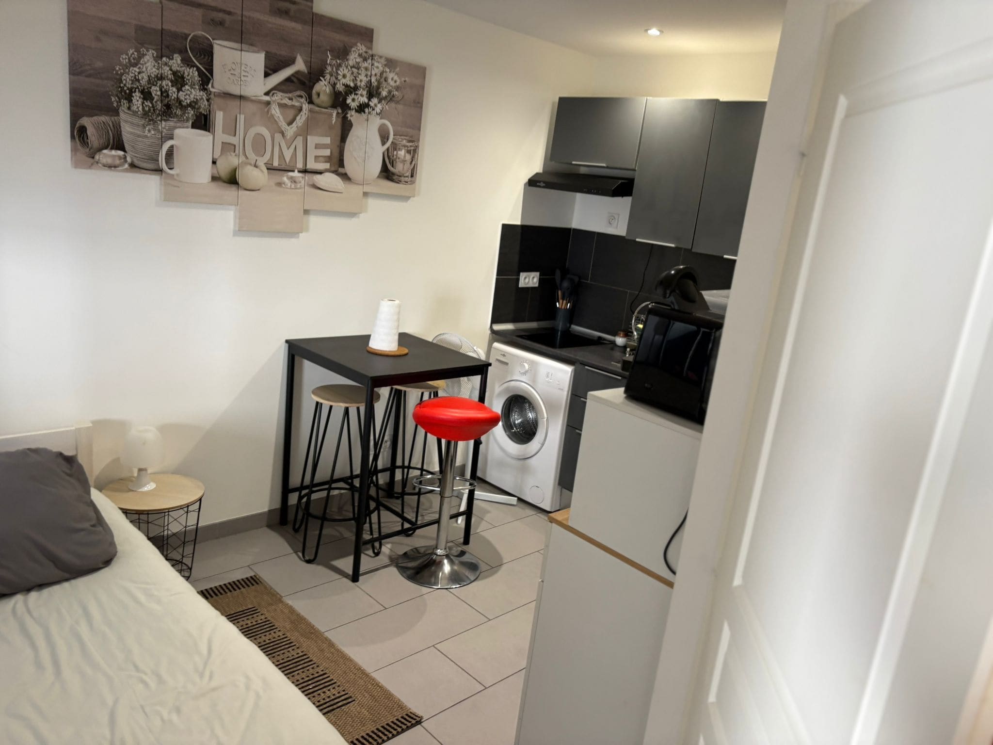 A compact living space with a bed, a small dining area with a table and two stools, a washing machine, and a kitchenette with cabinets and a microwave.