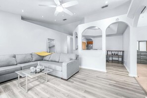 A modern living room with a grey sofa, a glass coffee table, and a ceiling fan. - Cottage on the Bay Pool Home (Tampa)