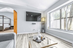 A modern living room with a flat-screen TV, a glass coffee table with decorative items, a sofa, and a window with blinds. - Cottage on the Bay Pool Home (Tampa)