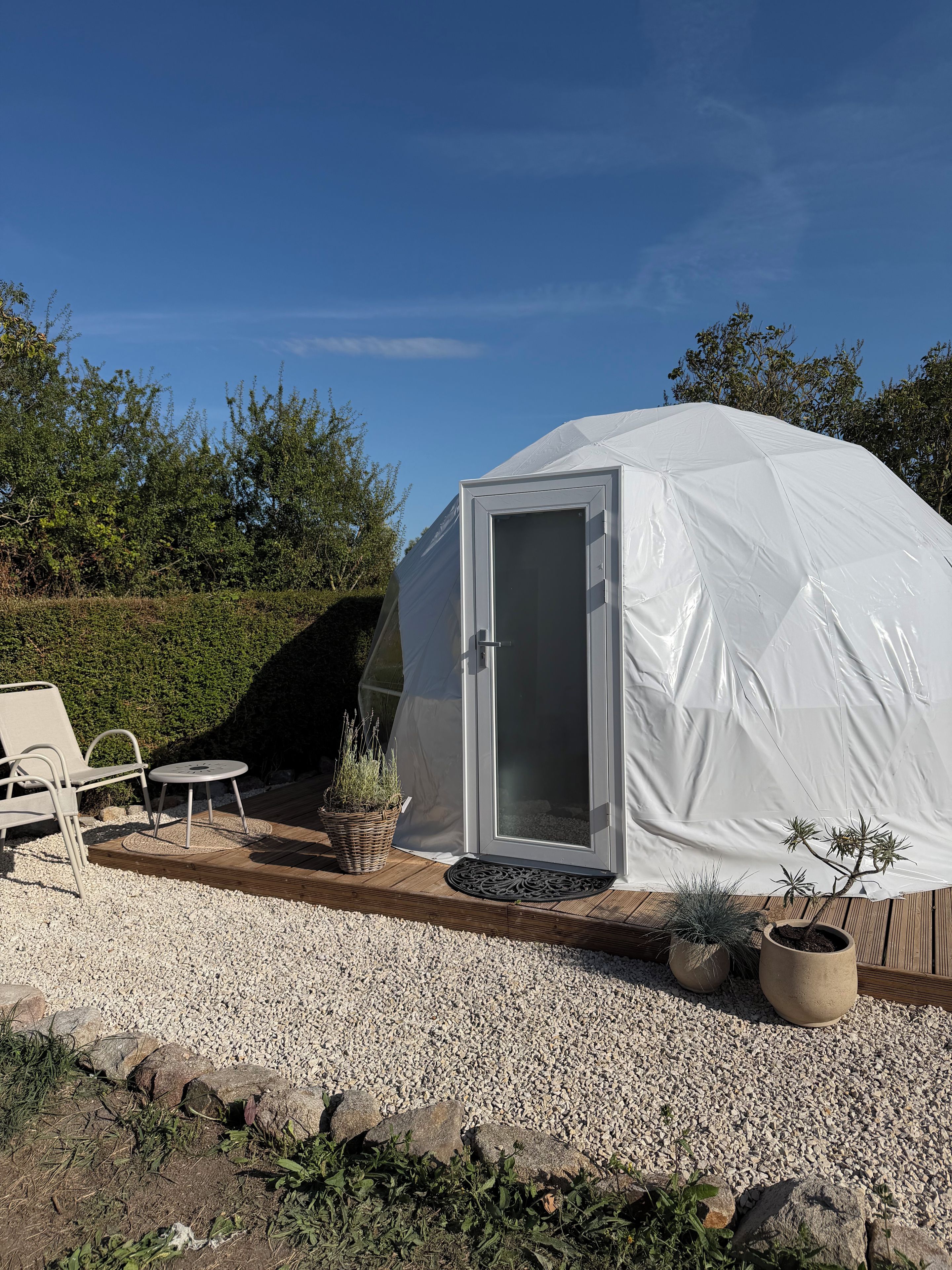 A white tent with a door, surrounded by a gravel area with potted plants and outdoor furniture.