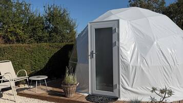 A white tent with a door, surrounded by a gravel area with potted plants and outdoor furniture.