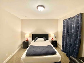 Une chambre d’hôtel avec un lit simple, deux tables de chevet, une petite table, un luminaire au plafond et un rideau foncé.