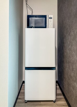 A white refrigerator with a microwave on top, placed in a corner with a wooden floor and a plain wall. - HANASTAY HIRO (Osaka)