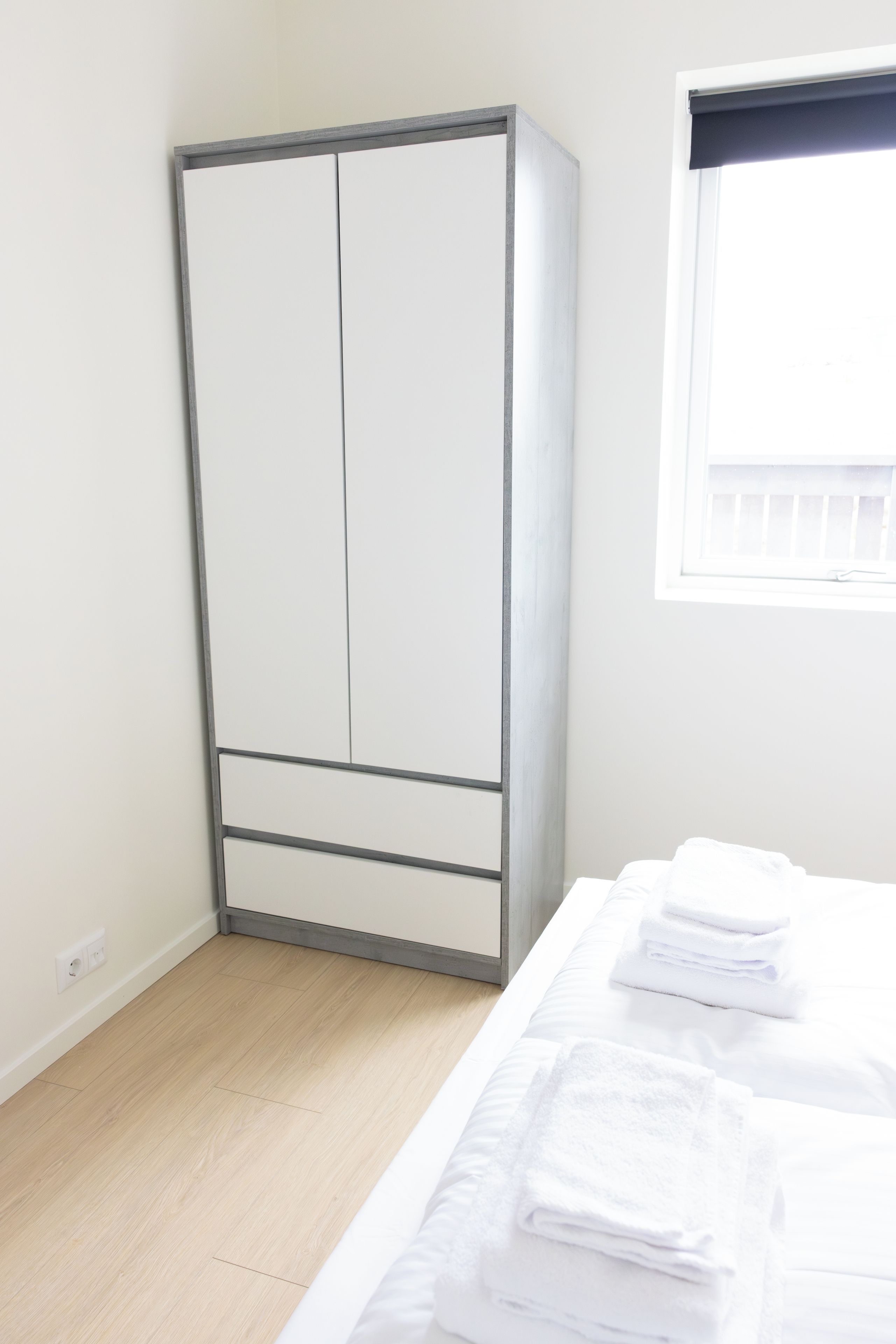 A bedroom with a wardrobe, a bed with white linens, and a window with a white blind.