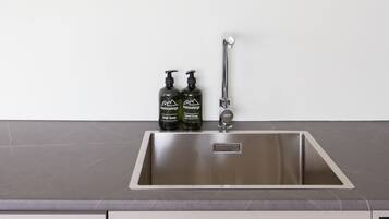 A kitchen with a stainless steel sink, two bottles of dish soap, and a potted plant on a wooden shelf.